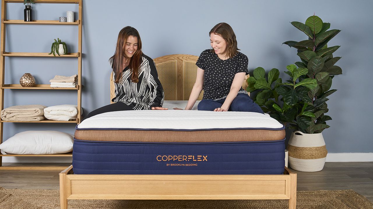Two girls sitting on navy, bronze and white Brooklyn Bedding CopperFlex Hybrid Pro mattress in sleep studio with rattan bedframe, shelf and green plants