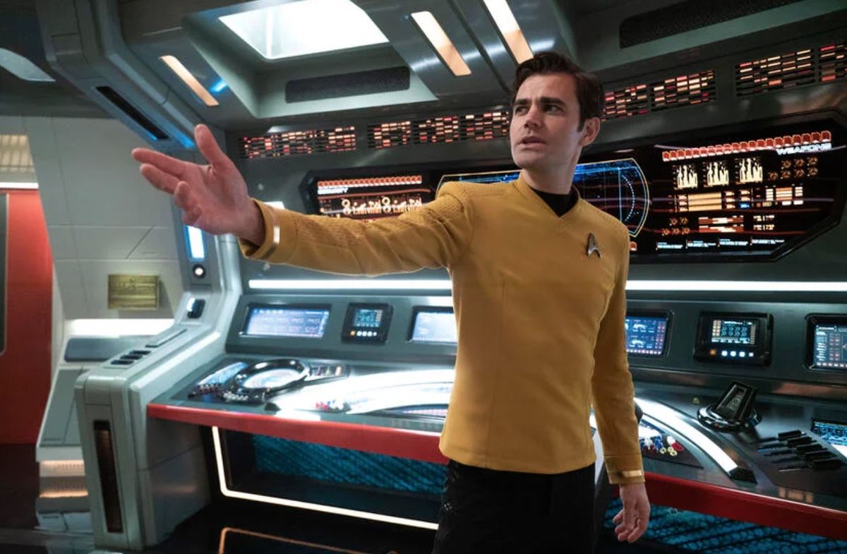 A man in a yellow Star Trek command uniform sings with arm outstretched. 