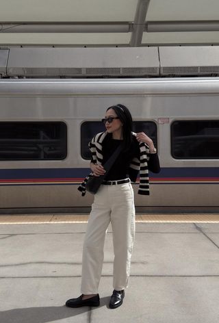 A winter travel outfit is shown in a photo of a woman standing in front of a train wearing a black boat neck top with a black belt, white jeans, black loafers, a black and white striped cardigan, a black headband, a black crossbody bag, and sunglasses