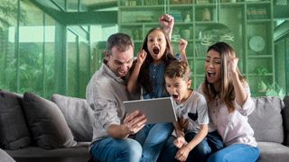 Young family sitting on a sofa and all looking excited while looking at a tablet.
