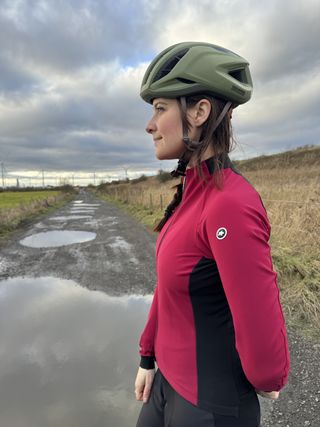 Women in pink and black jacket looks to the side, showing black side panels and sleeves. In front of muddy field.
