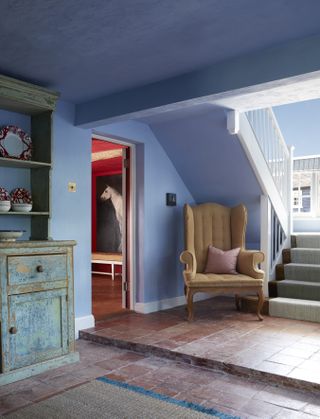 blue painted entryway with terracotta tiles, staircase, vintage armchair, sideboard with paint effect, red living room view through doorway