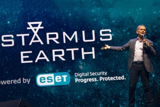 A man stands holding a microphone in one hand in front of a large display with the words starmus earth written in large letters.