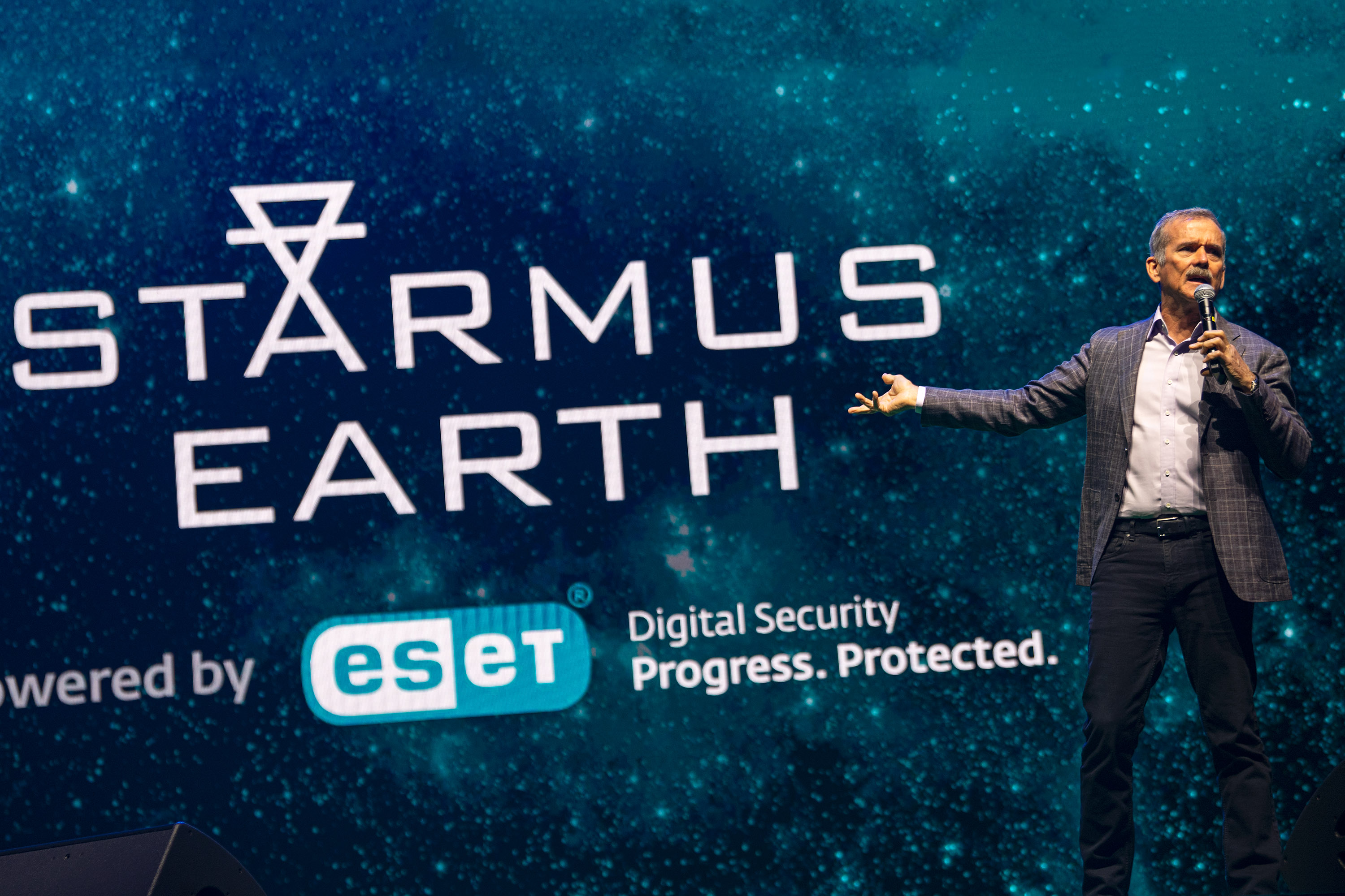 Un hombre está de pie sosteniendo un micrófono en una mano frente a una gran pantalla con las palabras Starmus Earth escritas en letras grandes.