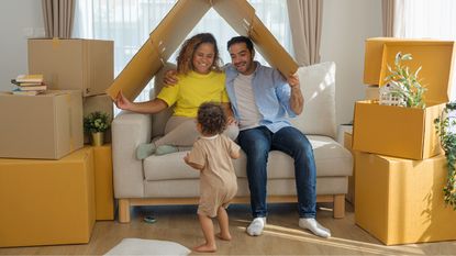 Young parents hold a folded cardboard box over their heads while playing with their toddler in the living room.