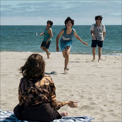 a woman (sofia vergara as griselda) sitting on a blue striped beach towel with her back to the camera faces three boys running towards her on the sand, with the sea in the background