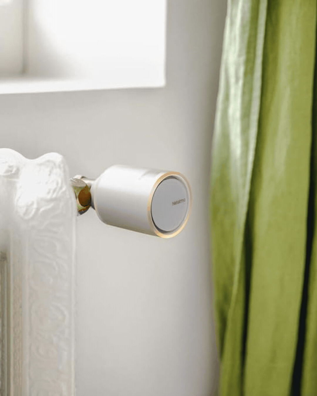 Netatmo Pack of 3 Connected and Smart Thermostatic Heads, seen as installed on a radiator and as a set next to a phone and placed against a dark green background.