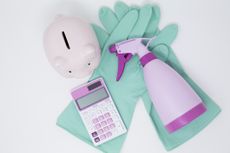 cleaning products in pastel colors, including a pink spray bottle and calculator and teal gloves, next to a light pink piggy bank