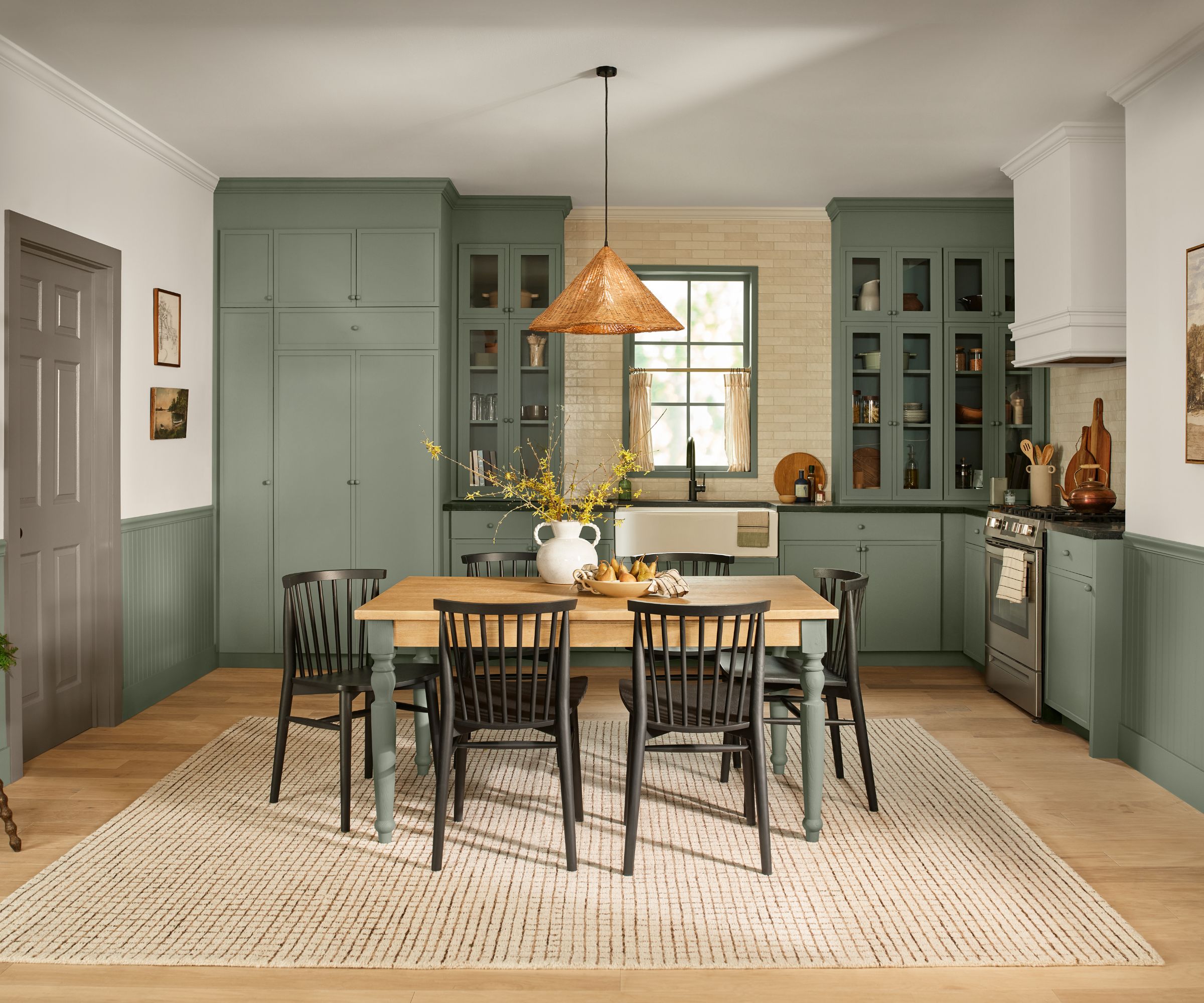 Kitchen with green painted cabinetry and cupboards, wooden floor with jute rug and a wooden dining table with rattan light