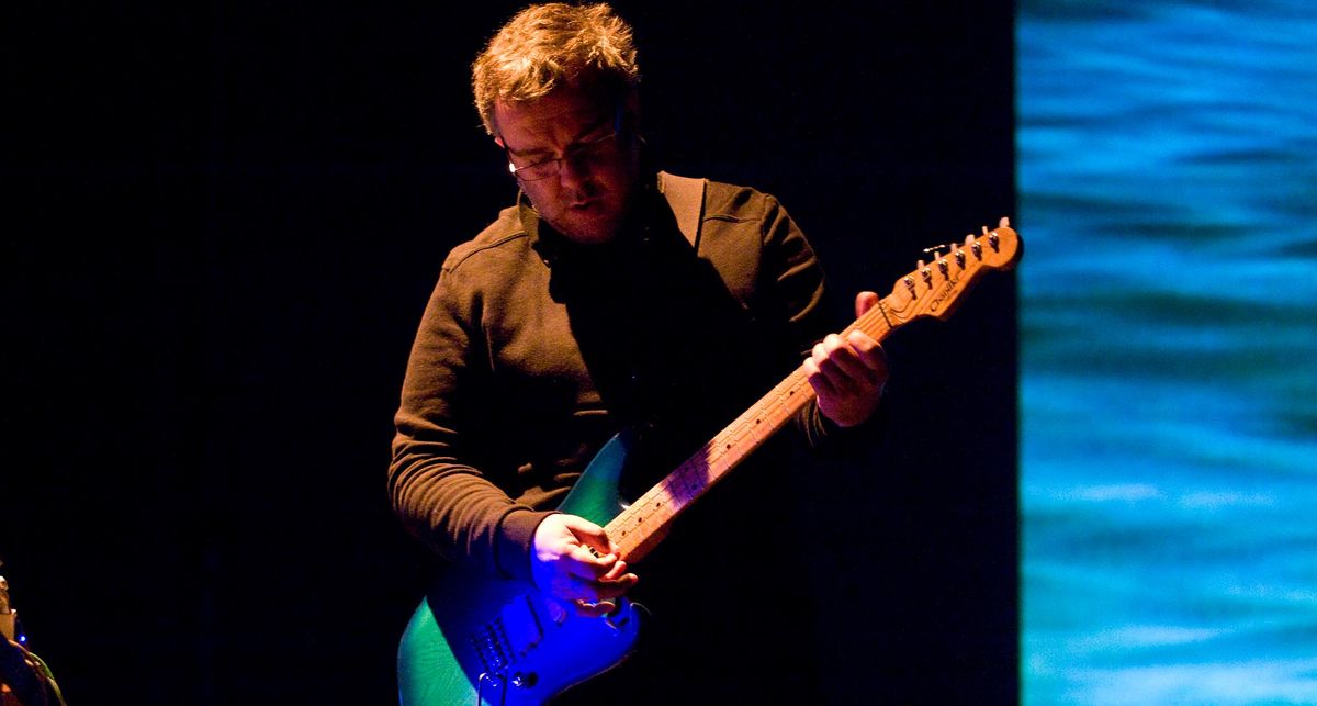 Robin Guthrie plays a blue Jazzmaster onstage, with a backdrop of the ocean complementing the guitar&#039;s finish. 