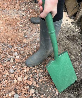 A person wearing khaki green wellington boots using a green shovel in backyard