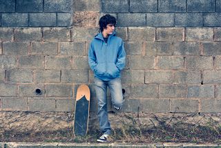 A teen boy stands against a wall, alone.