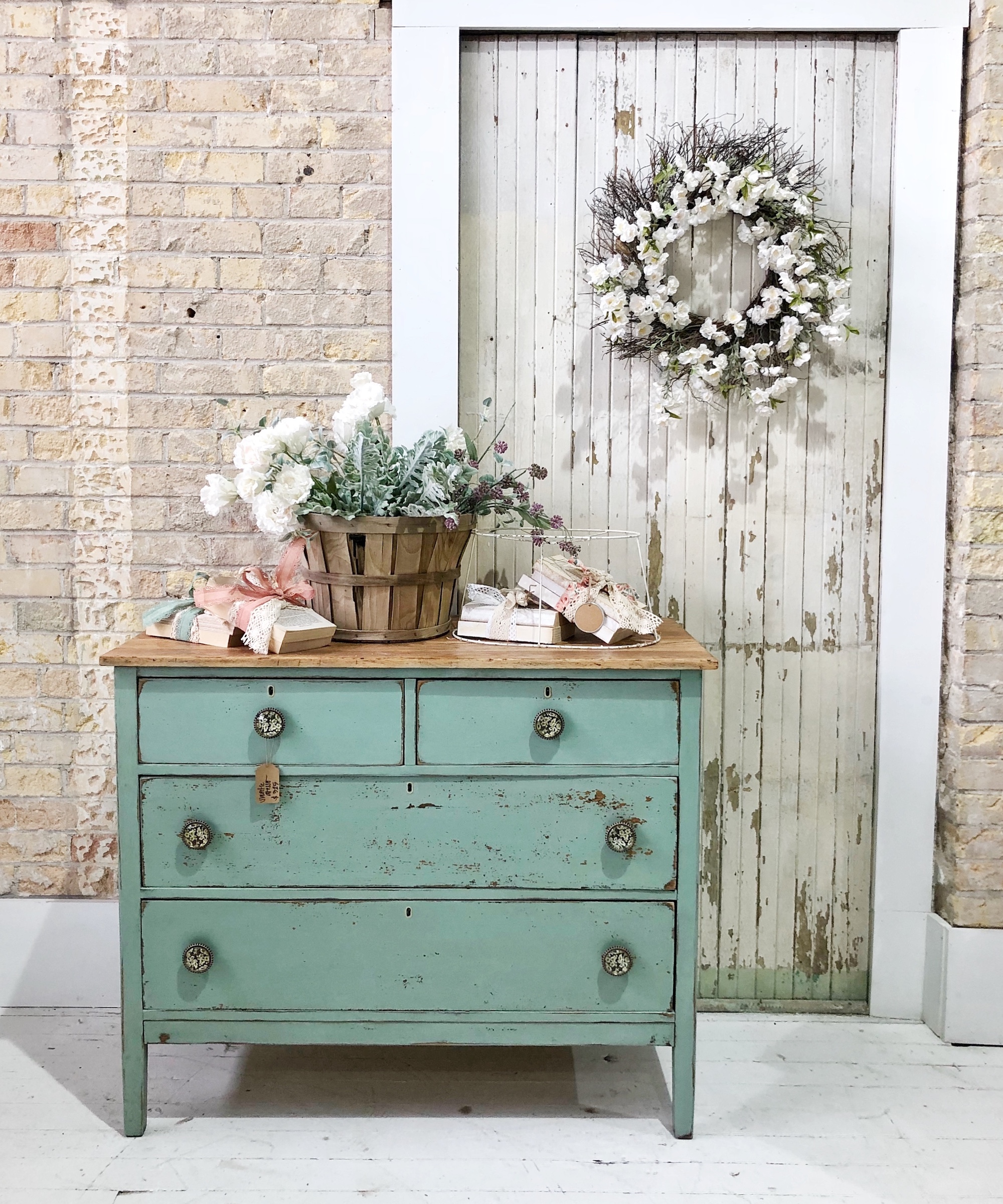 Chest of drawers with blue distressed finish created by using milk paint with decorative accessories above and brick wall and distressed wood door behind