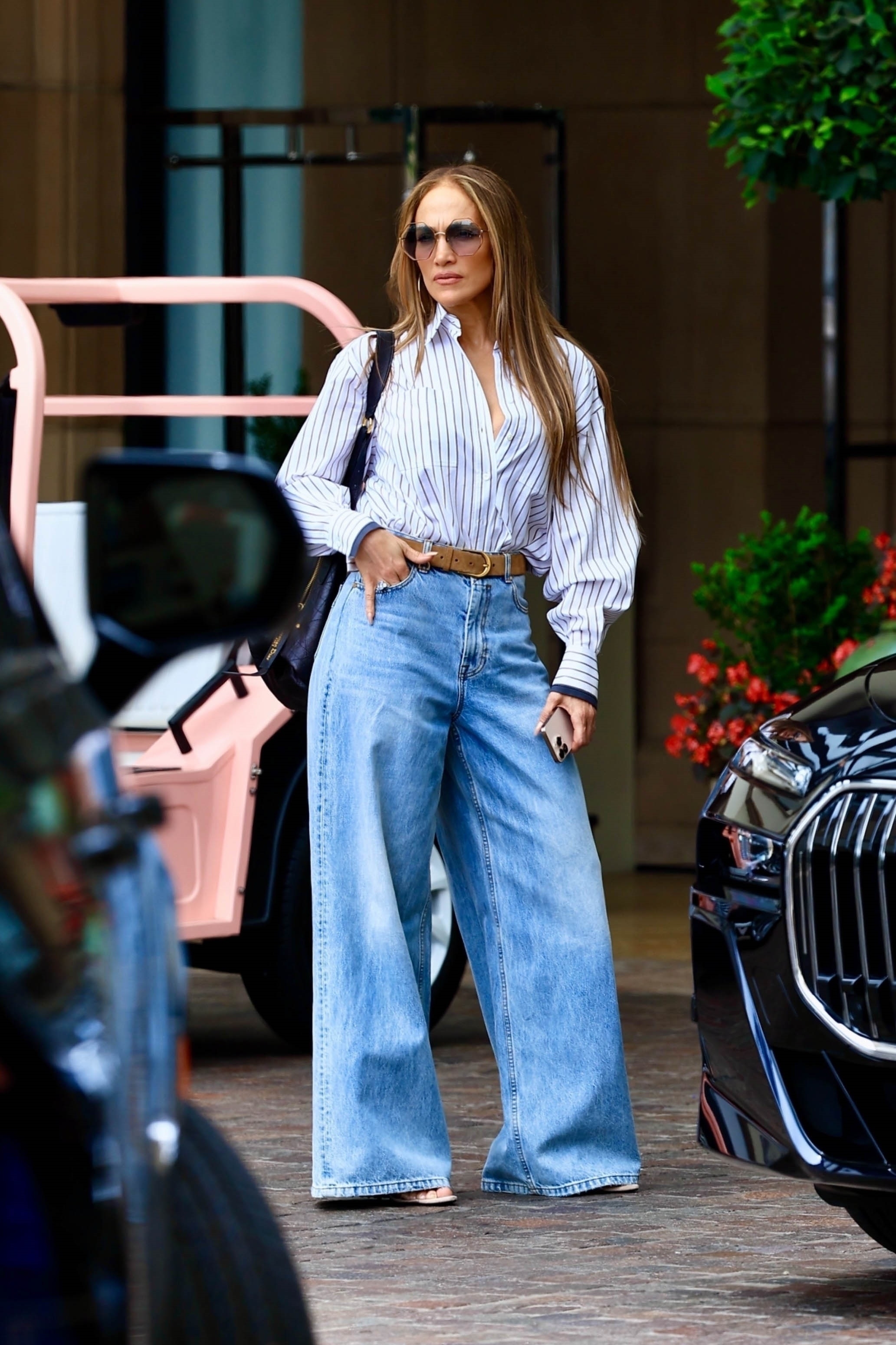 J.Lo wears a stripe button-down shirt, brown belt, wide-leg jeans, and a black shoulder bag.