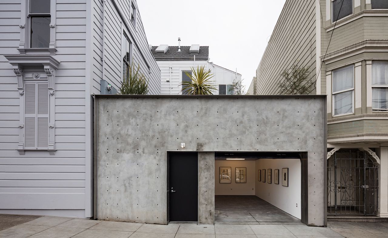 The former home of David Ireland photographed from the outside. Concrete rectangle-shaped structure with a black front door. The garage door is open and we see pictures in black frames hung on the wall.