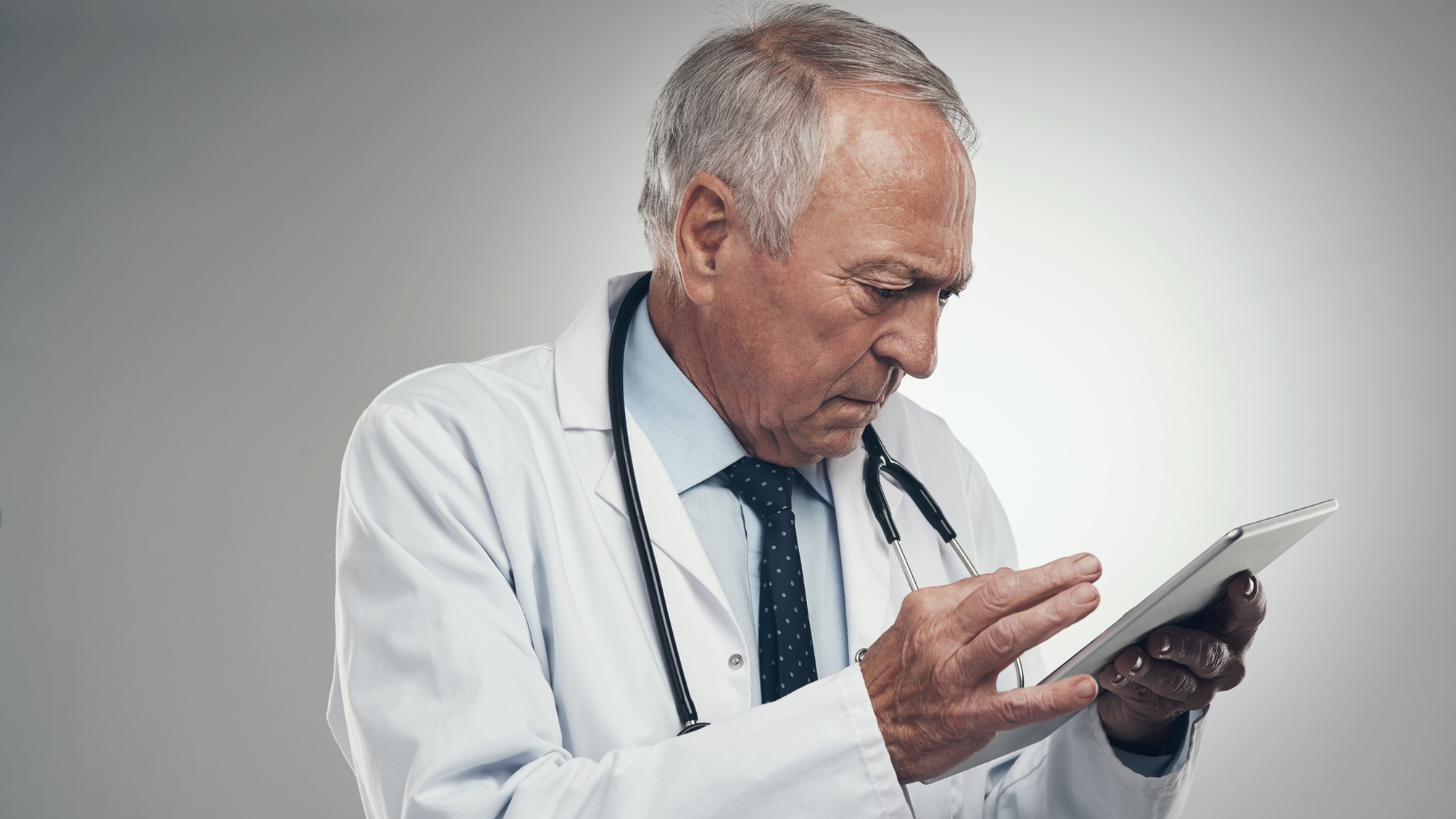 An older doctor looks intently at a tablet.
