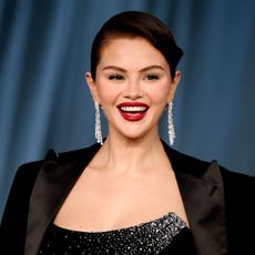 a woman with dark hair wearing a black blazer and an updo posing for paparazzi in front of a blue background