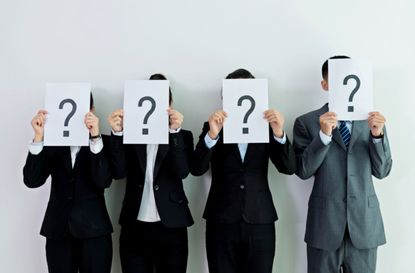 Four people in business suits hold up papers with questions marks on them in front of their faces.