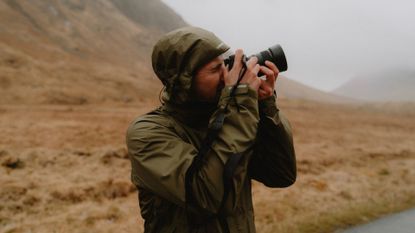 Male photographer in hooded jacket using camera below mountain, Glen Etive, Scottish Highlands, Scotland