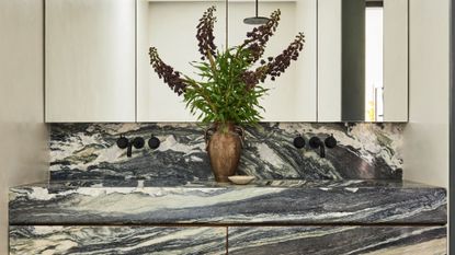 A marble his and hers sink with a bronze vase of flowers, black hardware on the backsplash and mirrored cabinets