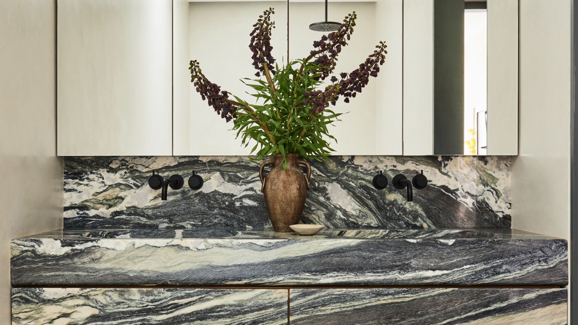 A marble his and hers sink with a bronze vase of flowers, black hardware on the backsplash and mirrored cabinets