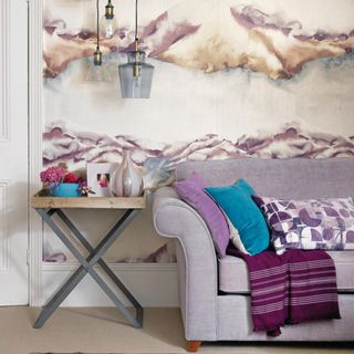 Patterned wallpaper, abstract cloud patterns in a living room with purple sofa, cream carpet, pendant lights and throws.