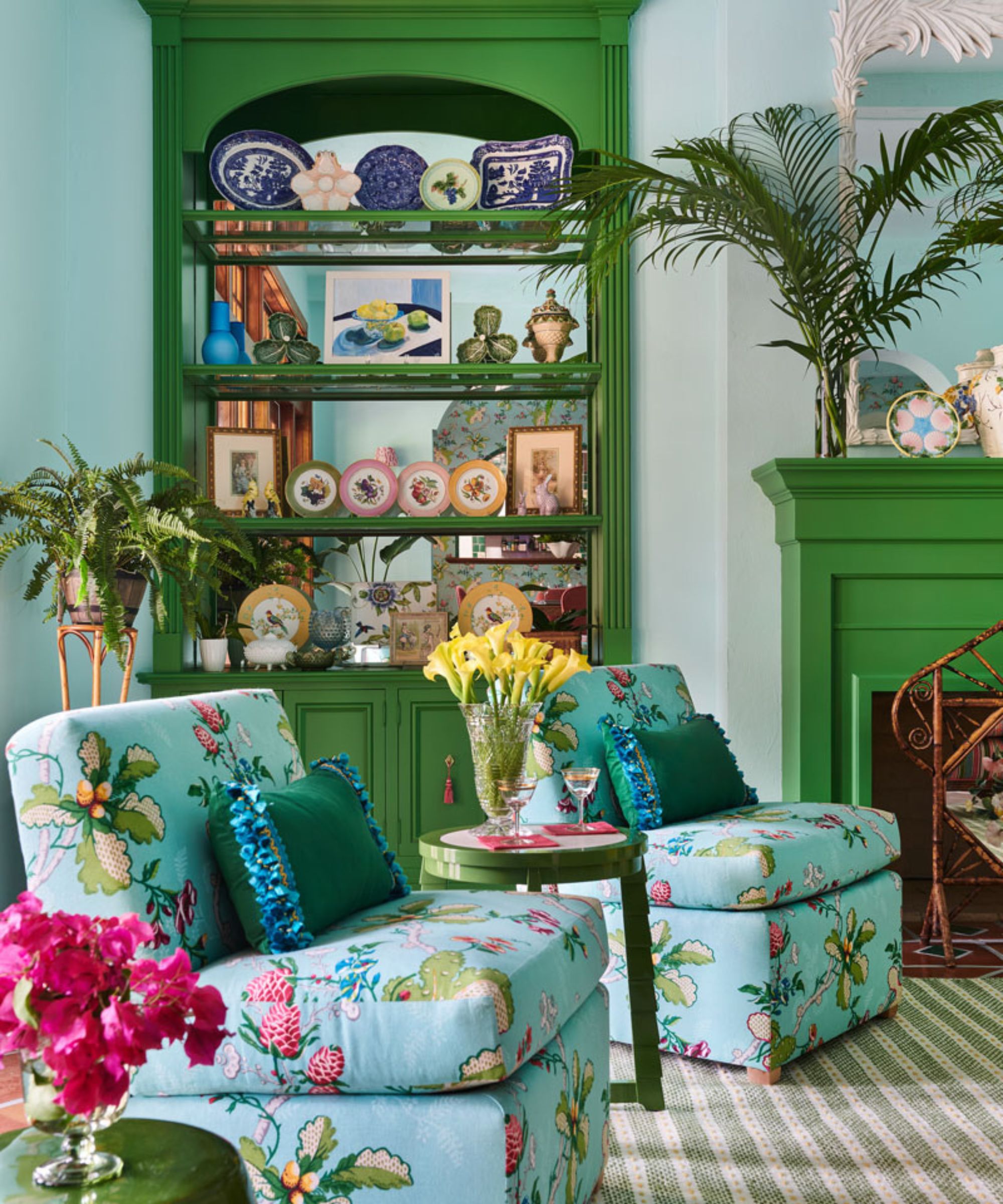 Living room with pale blue walls, a built-in cabinet with a mirrored back and plates sat on shelves, a green fireplace, and two armchairs upholstered in a blue floral fabric on a striped carpet