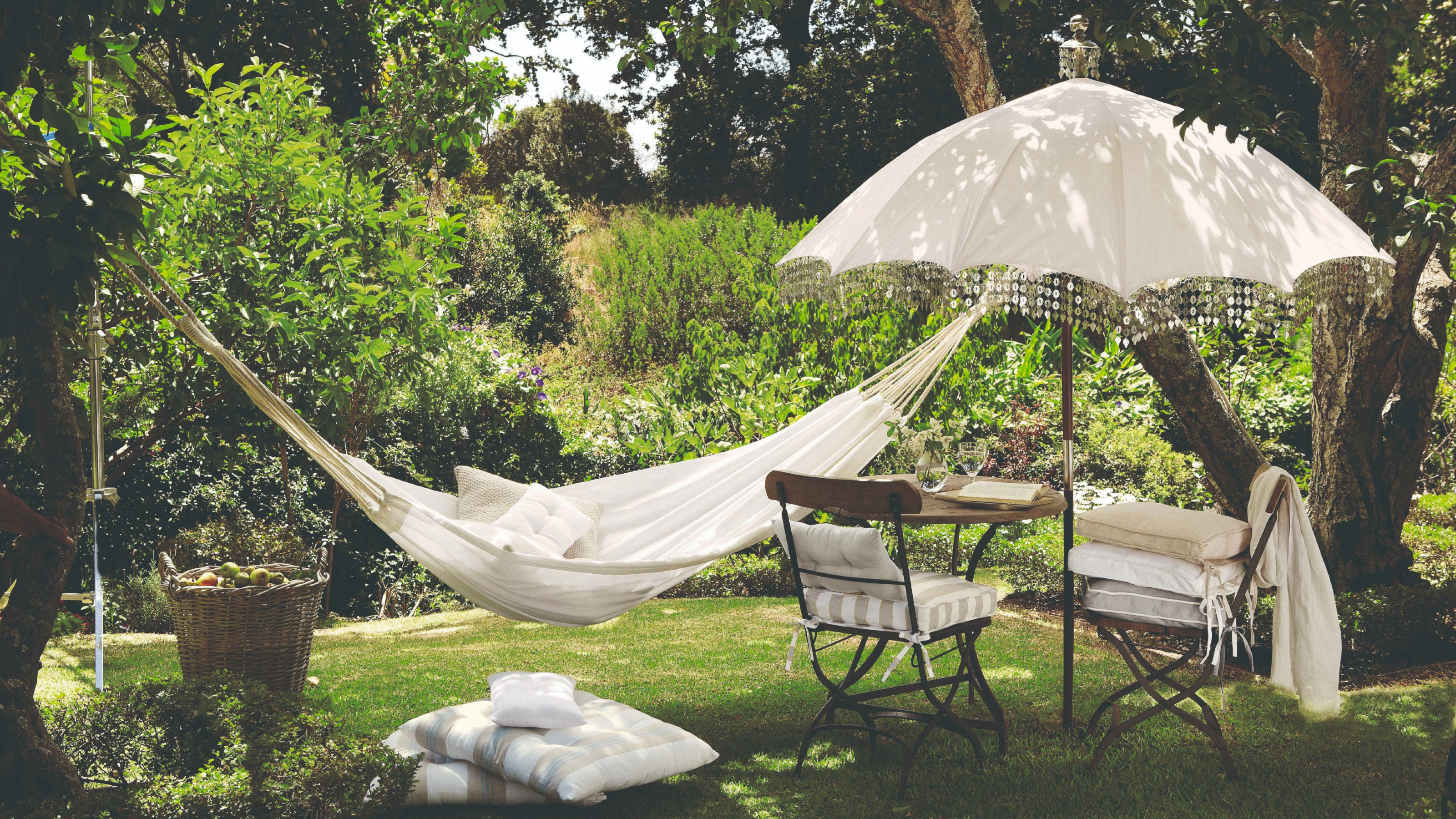 An English garden with a fringe-trimmed white parasol, a bistro set underneath it and a white hammock tied to the tree next to it