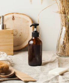 A dark brown glass soap dispenser on top of a beige cloth, with a wooden board and dried grass behind it