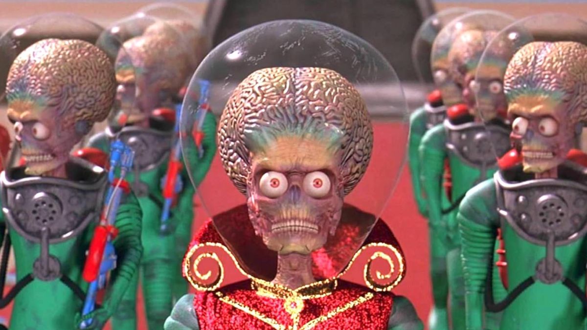 Screenshot from the movie Mars Attacks! A red-caped wearing alien with a big bulging brain protected by a big glass helmet is walking down a red carpet. They are flanked on either side with 3 other big-brained aliens in green uniforms.