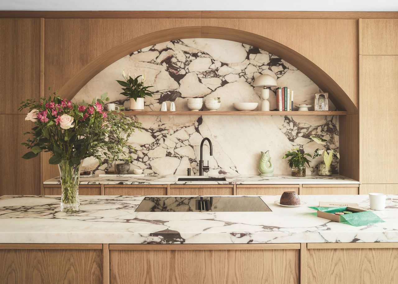 A modern marble and wood kitchen with an arched wood design above a marble backsplash with open shelving and a marble island with a vase of flowers and a hob on top