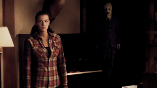Liv Tyler stands alone in a living room and a mysterious masked figure looms behind her