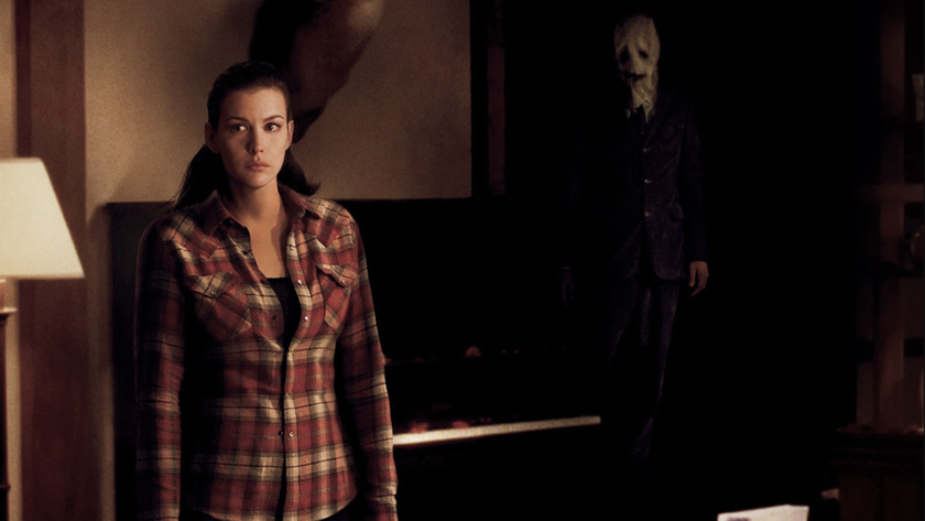Liv Tyler stands alone in a living room and a mysterious masked figure looms behind her