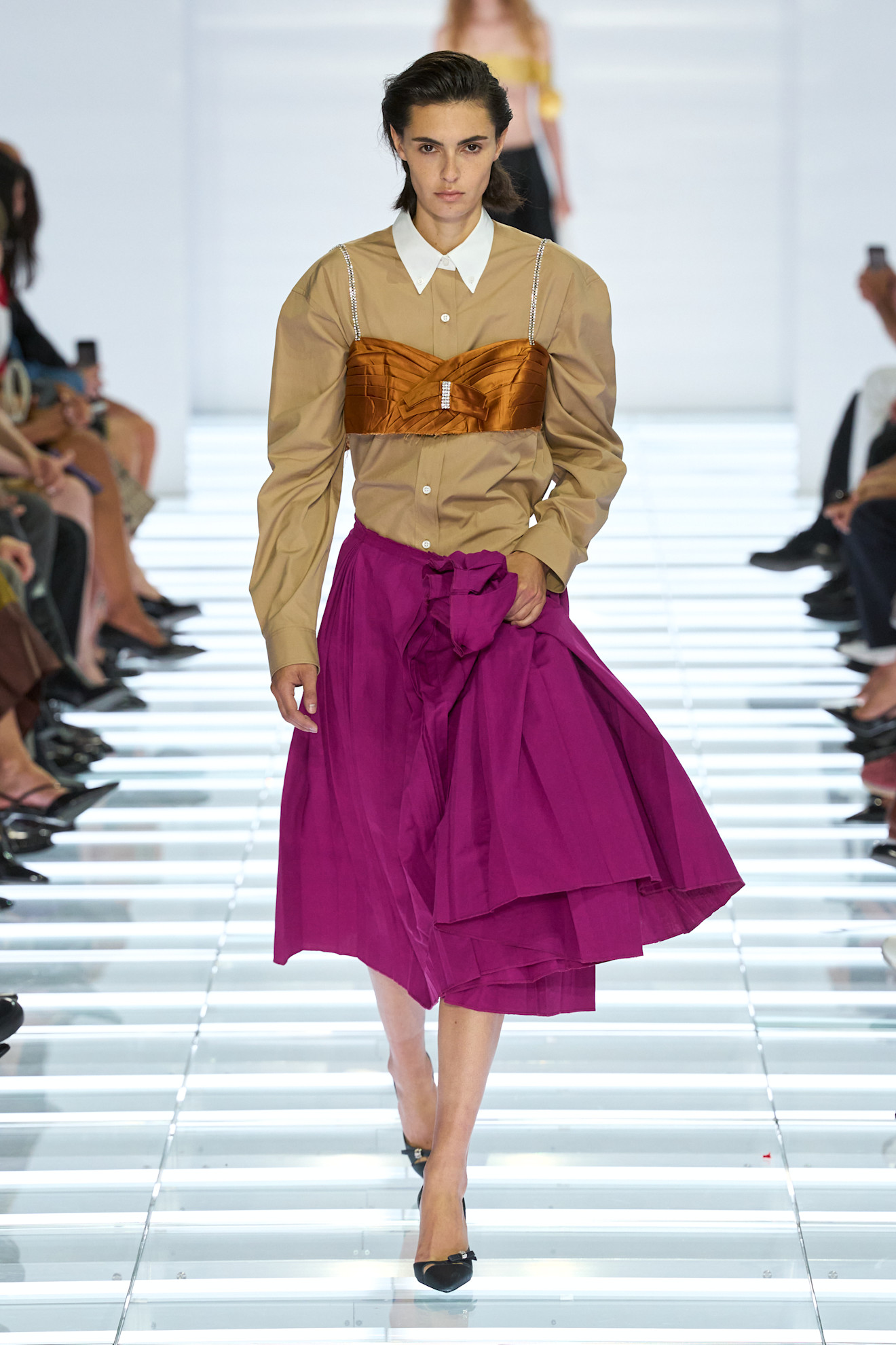 Model walks the Prada Spring 2027 runway in a magenta full skirt, tan button-down shirt, and bronze embellished bra top.
