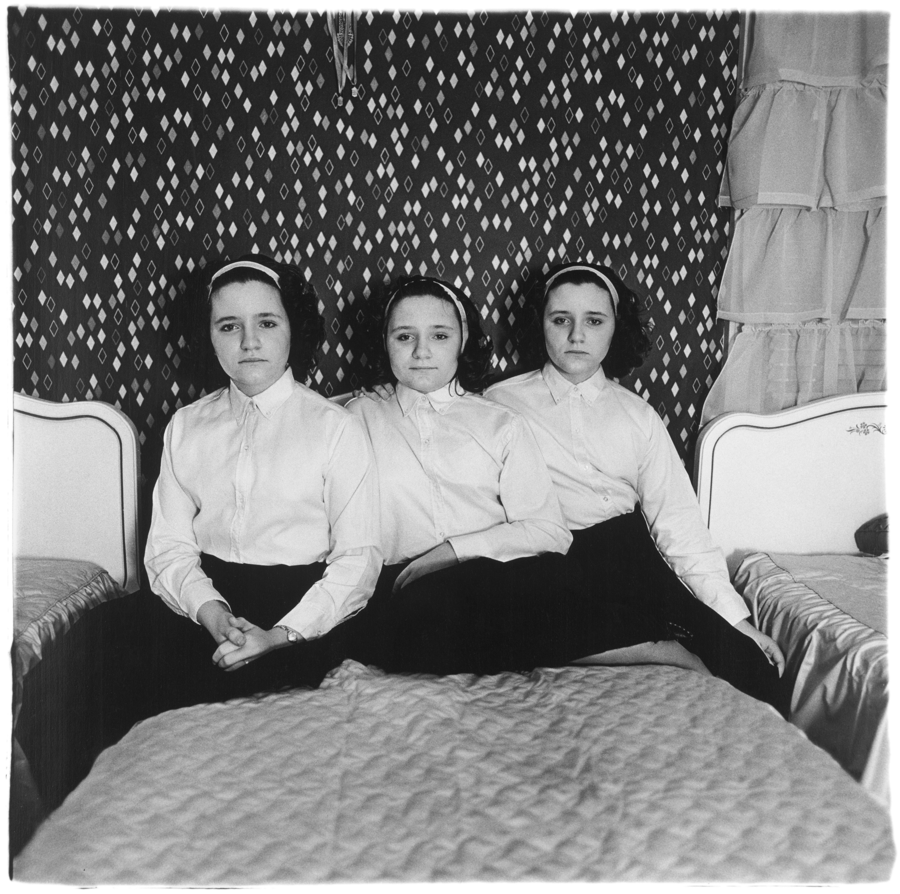 Three young, identically dressed triplet girls with dark curly hair and headbands sit closely together on a bed, staring directly into the camera in a room with a diamond-patterned wallpaper.