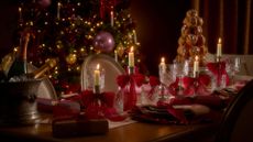 A Christmas tablescape with white tapers on brass holders with red ribbon bows by a croquembouche tower and an ice bucket of champagne by crystal coupes