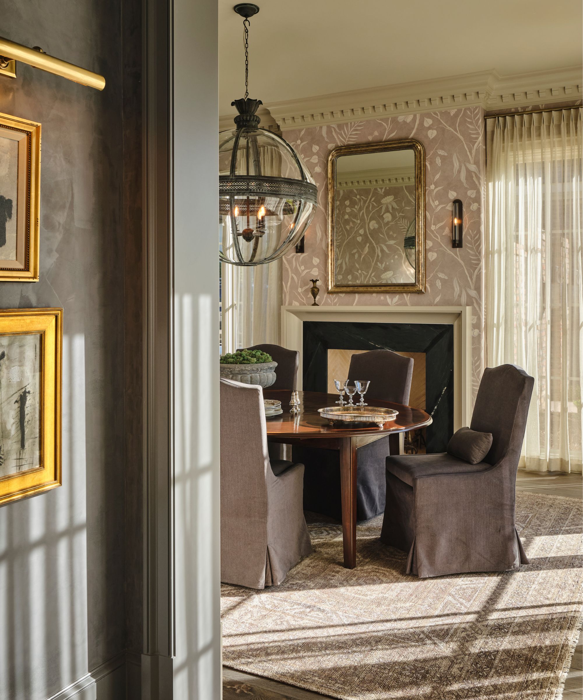 a soft neutral and mauve dining room with floral wallpaper, soft drapes, a fireplace, and linen slipcovered chairs around and antique table