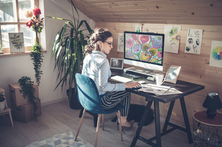 An artist sits at her desk with her traditional and digital art around her. 