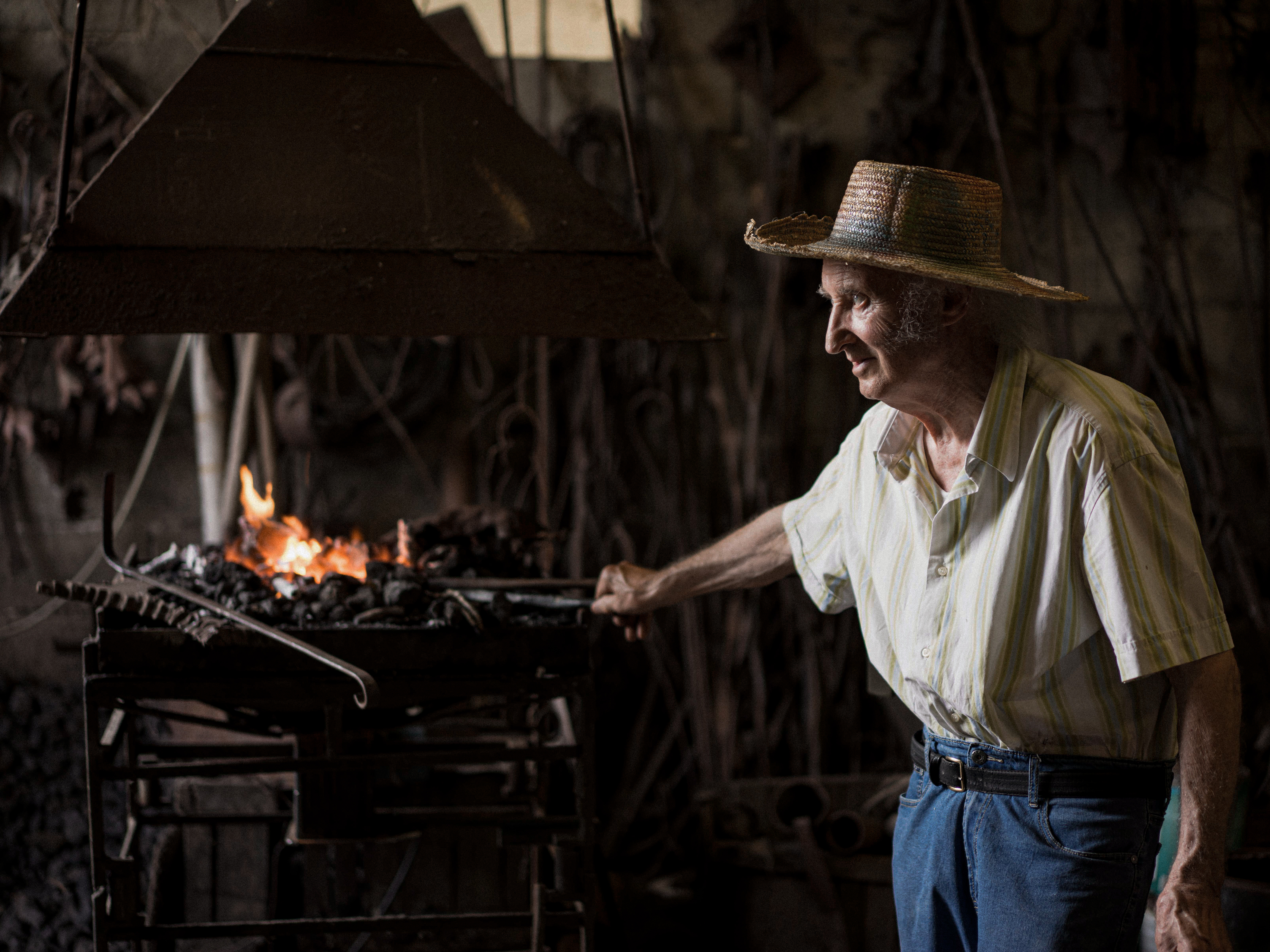 An old man dressed in a white and green, pale short-sleeved shirt, denim jeans, and a straw hat works in front of an iron oven on a metallic sculpture.
