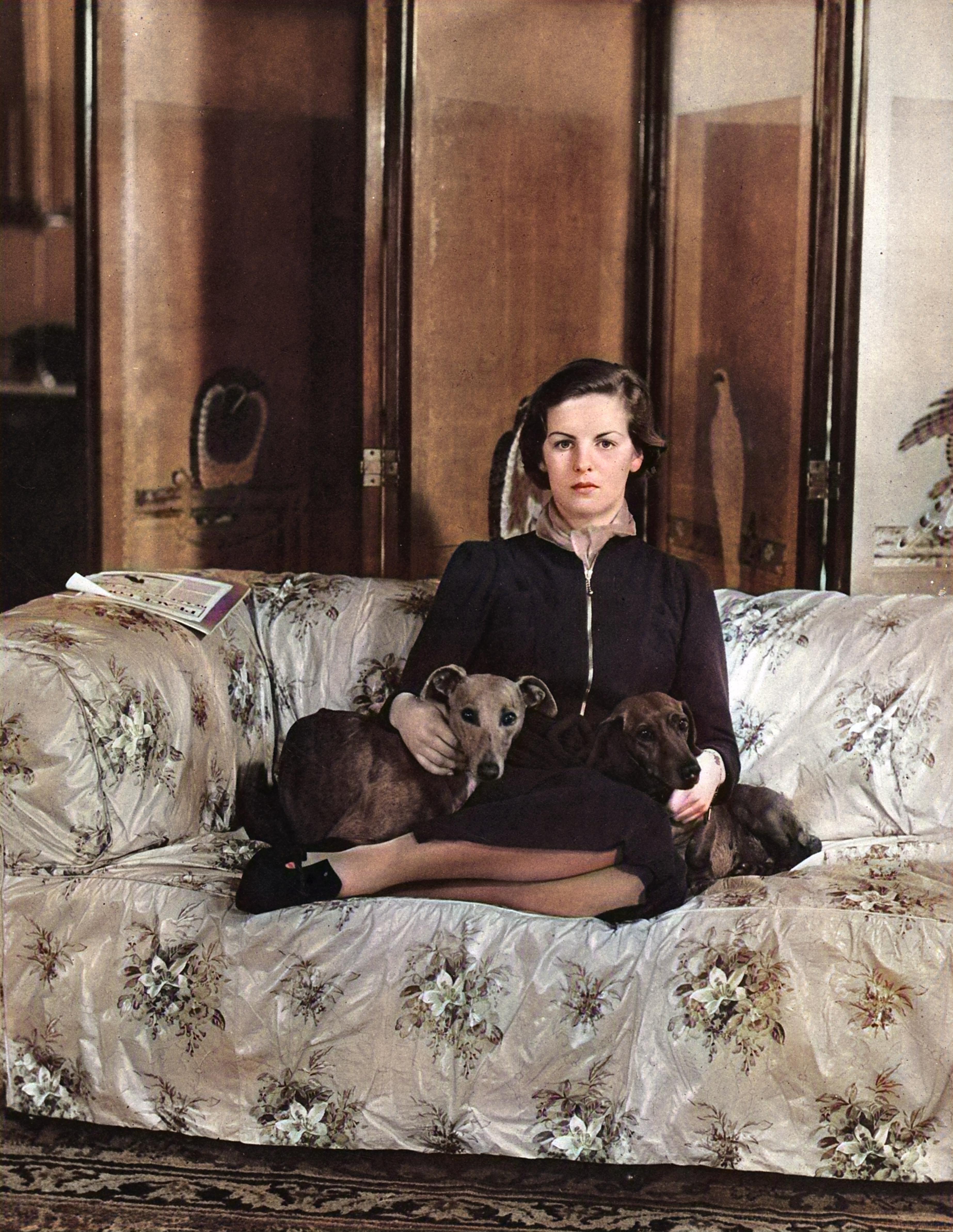 The Hon. Deborah Mitford, later Duchess of Devonshire. Pictured in the year she was a debutante — 1938 — on a floral sofa with two of her dogs, a greyhound and a dachshund.