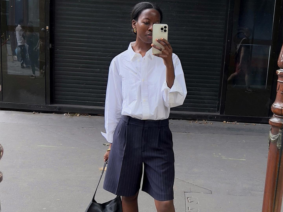 A woman wearing a white button up and blue long line shorts.