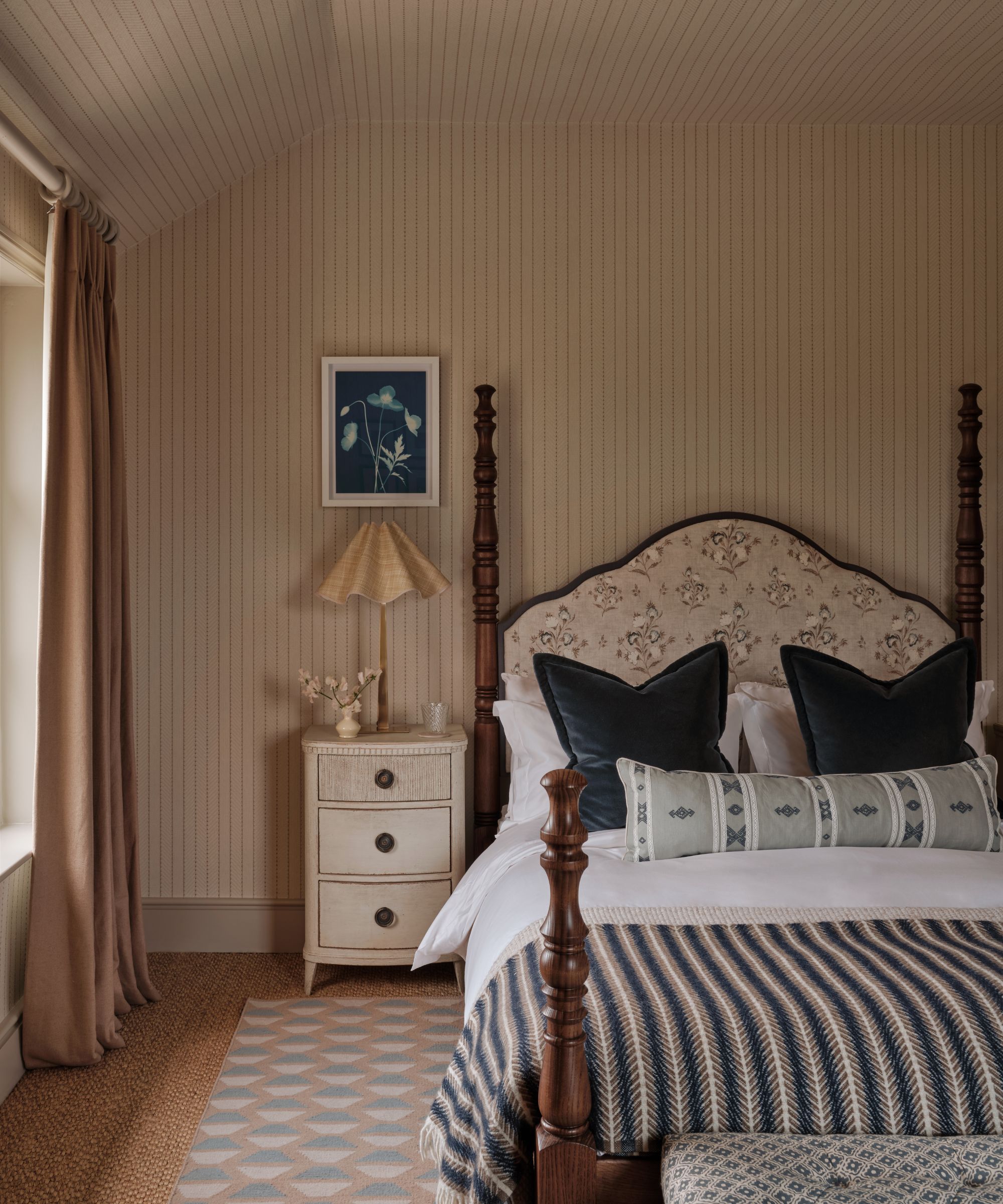 Cozy bedroom featuring a four-poster wooden bed with an upholstered headboard, fine striped wallpaper, a white nightstand with a fluted lampshade, and a patterned blue area rug.