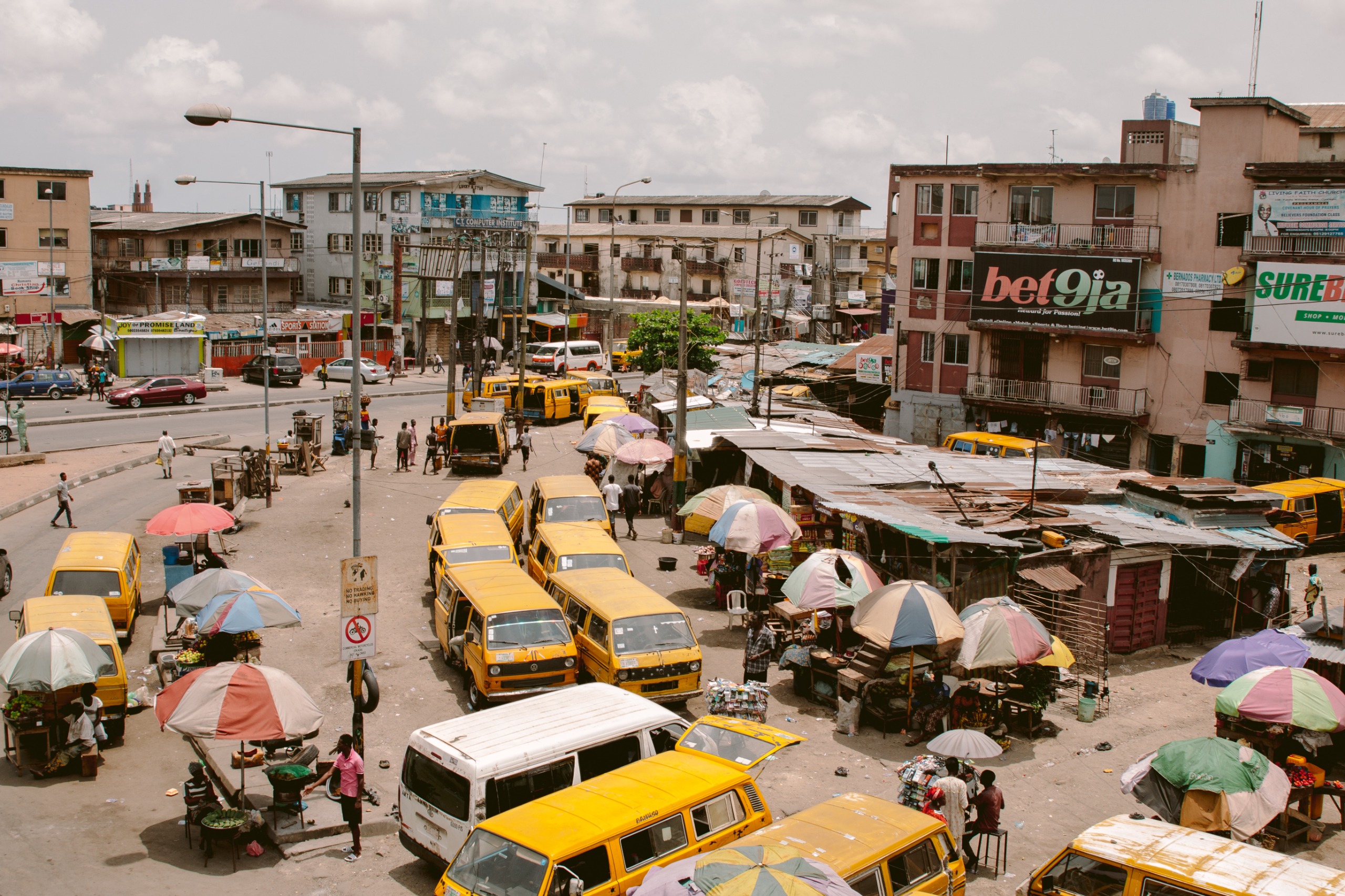 An aerial view of a busy Lagos bus park, with rows of yellow minibuses, market stalls and umbrellas packed along a dusty street.