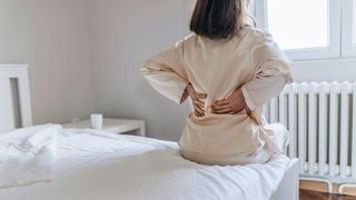 The back of a woman sat on the edge of her bed holding her lower back