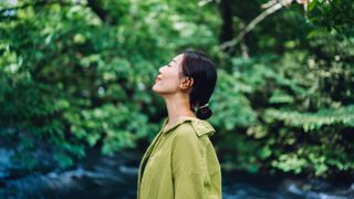 Woman in green blouse standing in nature with eyes closed smiling