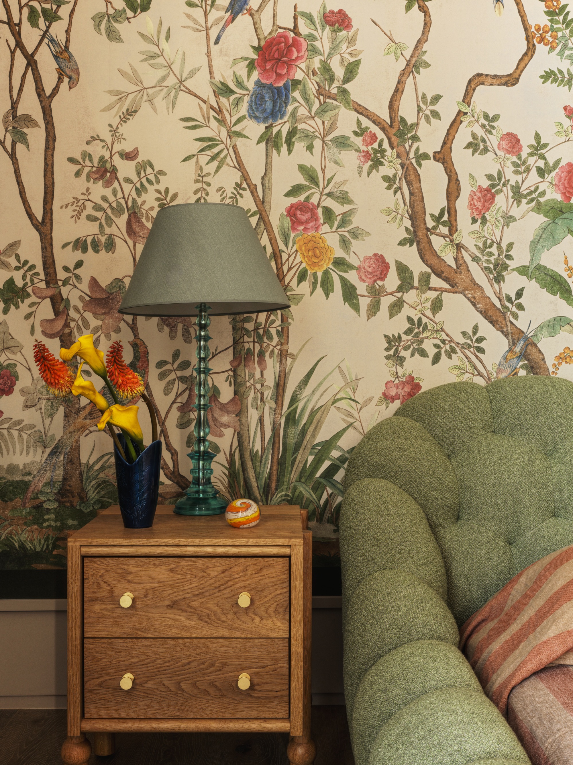 close up in bedroom with floral wallpaper, green bedframe, timber side table, green lamp with vase of flowers