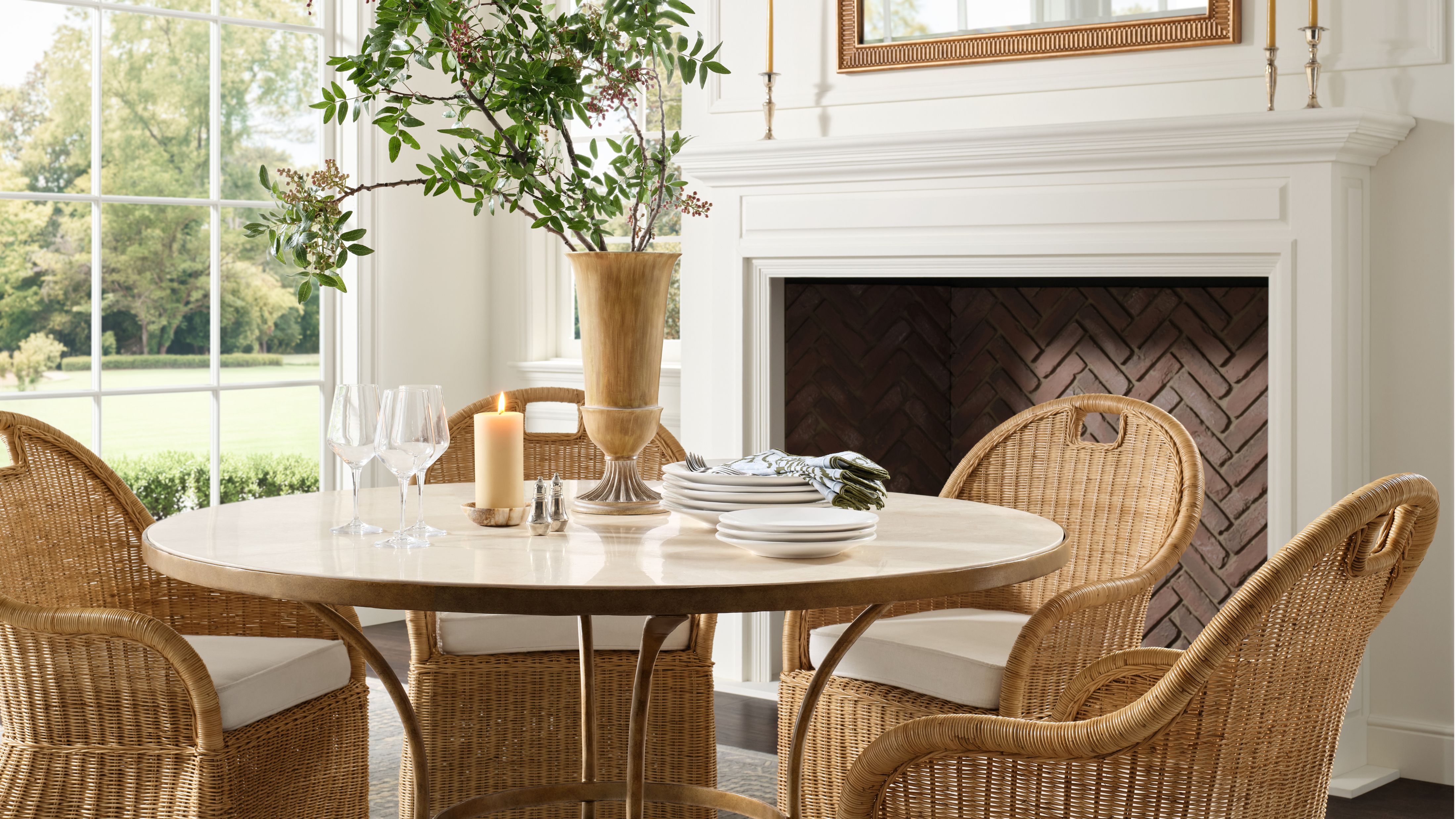Light-filled dining room area with a herringbone brick fireplace styled with a metal-and-stone circular dining table, woven dining chairs, and brass tabletop accents