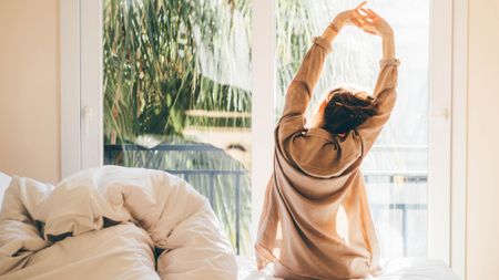 A well-rested woman with dark hair piled up in a messy bun sits on the edge of her bed and stretches her arms up while facing a sunny window early in the morning