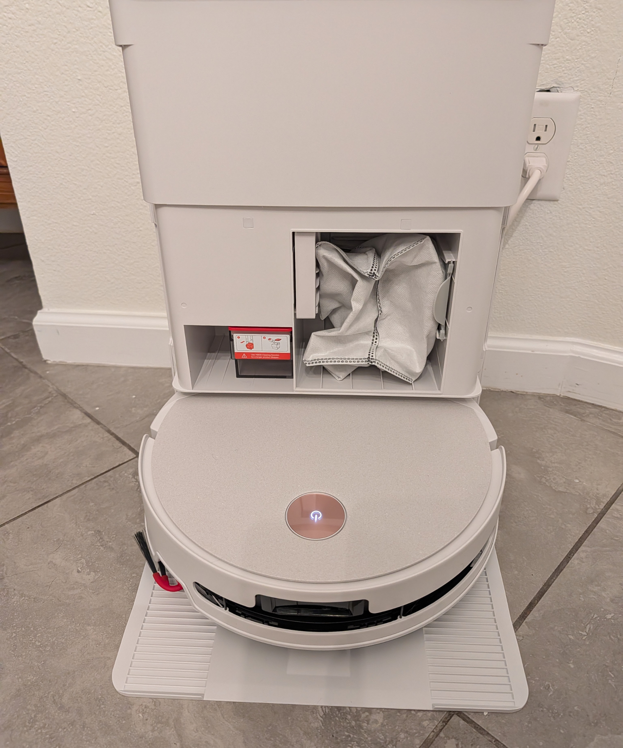 Yeedi M16 Ultra in dock with cover off showing dust bag and cleaning solution bin on a tile floor agains a white wall.