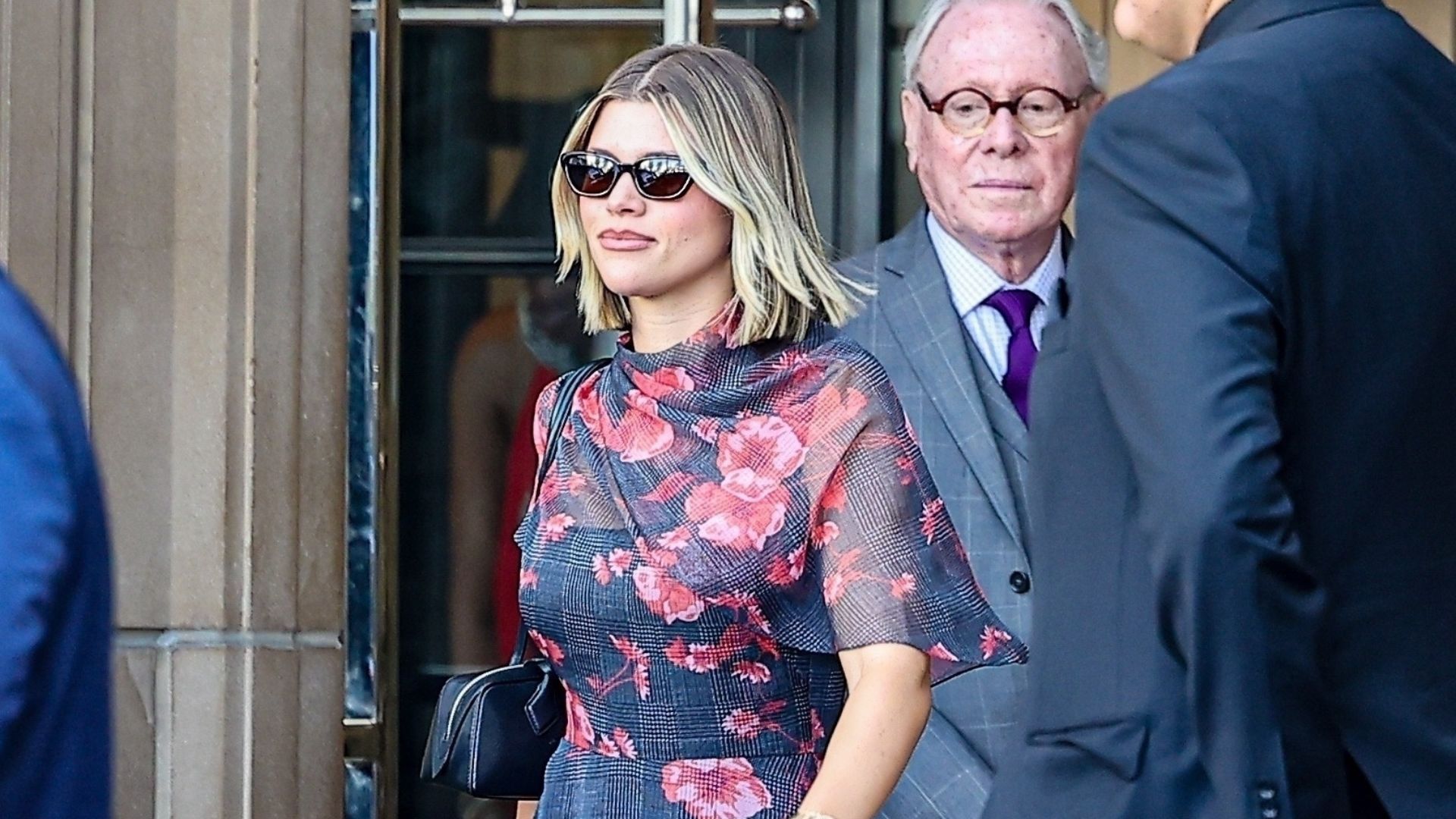 Sofia Richie wears a navy dress with pink floral embellishments with backless wedge mules and a black east-west bag.